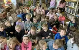 Liczna grupa dzieci siedzi na dywanie w bibliotece. Z prawej strony z boku na krzesłach siedzą opiekunowie.