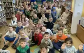 Duża grupa dzieci siedzi na dywanie, za nimi z tyłu stoi dw&oacute;ch aktor&oacute;w w niebieskich kombinezonach i czapeczkach z daszkiem.