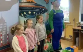 Z przodu stoją trzy dziewczynki i aktorka w niebieskim kombinezonie i kolorowej czapce z daszkiem. Za nimi tło statku kosmicznego na ziemi i motyle.