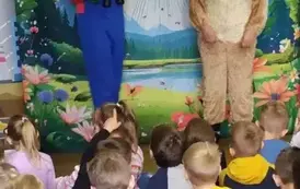 Przed dziećmi stoi przebrany piesek, a obok aktor w niebieskim kombinezonie, kt&oacute;ry trzyma notes i zielone okulary, na szyi wisi mu lornetka. W tle statek kosmiczny lądujący na ziemi.