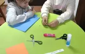 Dwie dziewczynki siedzą przy zielonym okrągłym stoliku na kt&oacute;rym są dwie kolorowe kartki, nożyczki, klej i pasek z wydrukowanym tekstem. Dziewczynka w ciemnych włosach ma przy sobie niebieską kartkę.