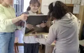 Dziewczynka w blond włosach ma włożone ręce w kartonie w kt&oacute;rym nie widać co jest w środku. Trzyma coś papierowego, przy niej stoi Pani prowadząca i Pani nauczycielka.