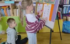 Dziewczynka w blond włosach w kitku stoi przy metalowej tablicy i na przyczepionej kartce oł&oacute;wkiem łączy ze sobą w pary te same zwierzątka. Obok siedzi chłopiec i patrzy.