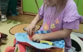 Dziewczynka w blond włosach siedzi na dywanie i bawi się pluszową książeczką.