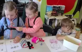 Dzieci przy stole wykonują karty pracy, jedna dziewczynka zagląda do drugiej  trzymając kredkę. Na stole leżą opr&oacute;cz kredek dwie pluszowe małe książeczki.