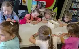 Dzieci siedzą przy białym stole i wykonują kredkami karty pracy.