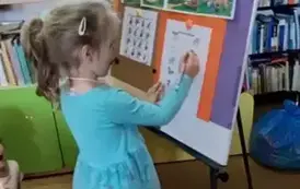 Dziewczynka w niebieskiej sukience stoi przy metalowej tablicy i na kartce oł&oacute;wkiem łączy takie same zwierzątka.