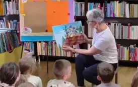 Pani sołtys siedzi na krzesełku bokiem do dzieci i z otwartą książeczką odwr&oacute;coną obrazkami do dzieci i czyta im bajkę.