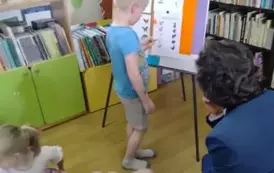 Dzieci siedzą na siedziskach, z przodu przy tablicy stoi chłopiec i uzupełnia kartę pracy. Sołtys siedzi obok i patrzy.