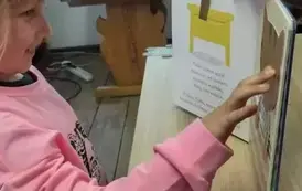 Dziewczynka w blond włosach i w r&oacute;żowej bluzce stoi przy biurku na kt&oacute;rym jest otwarta książka i palcem dotyka kotka, kt&oacute;ry jest uwypuklony w książce