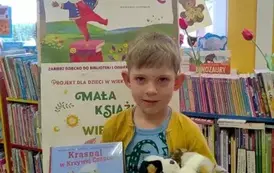 Chłopiec w blond włosach stoi przy tablicy z plakatem dotyczącym akcji Mała Książka Wielki Człowiek. Trzyma w ręku nową książkę i dwa pluszowe pieski.