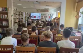 Cała bilbioteka jest wypełniona krzesłami, na kt&oacute;rych siedzą uczniowie SP, a z przodu szefowa wita wszystkich rozpoczynając wydarzenie. U g&oacute;ry jest ekran z wyświetlanym plakatem z wydarzenia.