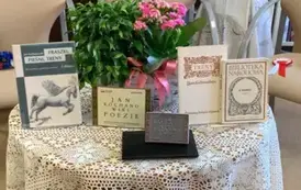 Na okrągłym stoliku jest obrus, kwiat w doniczce, postawione książki Jana Kochanowskiego i pieczątka z Narodowego Czytania