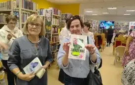 Z przodu widać dwie Panie kt&oacute;re trzymają zakupione książki, jedna z nich pokazuje okładkę. W tle widać ludzi rozmawiających ze sobą.