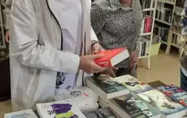 Przy wyłożonych książkach na biurku stoją dwie kobiety i patrzą. Jedna z nich trzyma książkę i czyta z tyłu opis. W tle widać inną kobietę w okularach.