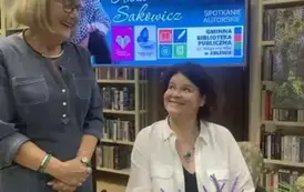 Pisarka siedzi przy stoliku i patrzy z uśmiechem na Czytelniczkę kt&oacute;ra stoi obok. Obie Panie bardzo uśmiechnięte. Nad nimi wyświetlony jest ekran z plakatem ze spotkania.