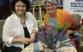 Uśmiechnięta czytelniczka przy stoliku przybliża się twarzą do pisarki trzymając książkę do podpisu. Obie Panie się uśmiechają. Za nimi jest w tle obraz starego domu z bzem do kt&oacute;rego przyczepione są grafiki okładek od książek pisarki.