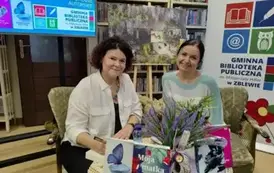 Przy stoliku pisarki siedzi czytelniczka prosząc o podpis na książce. Obie Panie uśmiechają się. Na stoliku są książki pisarki, wazonik z wrzosem i woda w butelce.