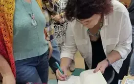 Na biurku są wyłożone książki pisarki, przy kt&oacute;rym atorka podpisuje książkę czytelnicze, kt&oacute;ra stoi obok. 