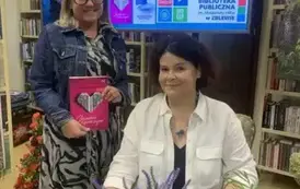 Anna Sakowicz siedzi przy stoliku, a przy Niej stoi czytelniczka trzymając książkę pisarki. Obie Panie uśmiechają się.