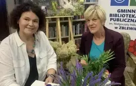 Pisarka przy stoliku podpisuje książkę czytelniczce. W tle widać rolap biblioteki, na środku jest obraz starego domu otoczonego bzem na kt&oacute;rym są przeczepione graiki okładek od książek pisarki.