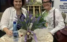 Przy okrągłym stoliku siedzą pisarka i jej czytelniczka, uśmiechają się. Na stoliku stoją : wazonik z wrzosem, sztuczna dynia, butelka z wodą i pudełko z kwiatami.