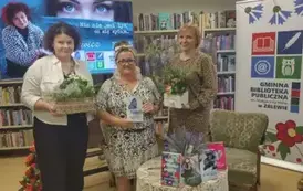 Przy okrągłym stoliku stoją Anna Sakowicz i Jej prowadząca z pudełkami kwiat&oacute;w, a po środku kierowniczka trzymając książkę "Niebieskie motyle" . 