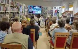 Wszystkie krzesła zajęte przez gości, z przodu widać pisarkę m&oacute;wiącą do mikrofonu. U g&oacute;ry jest duży ekran z plakatem ze spotkania.