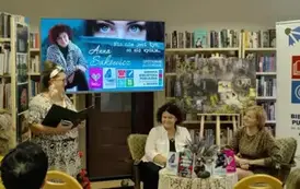 Pisarka i Jej prowadząca siedzą przy stoliku i się uśmiechają, a po lewej stoi kierowniczka biblioteki i wita wszystkich na spotkaniu. U g&oacute;ry jest duży ekran z wyświetlanym plakatem dot. spotkania.