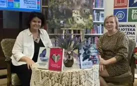 Przy okrągłym stoliku na kt&oacute;rym wystawione są książki pisarki siedzą Anna Sakowicz i Jej prowadząća spotkanie. Po prawej jest rolap biblioteki a po lewej u g&oacute;ry jest duży ekran z wyświetlanym plakatem dot. spotkania.