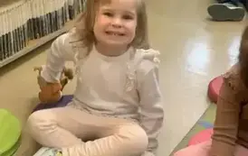 Dziewczynka w blond włosach bardzo się uśmiecha trzymając w ręce pluszową książeczkę. W tle widać inne dzieci.