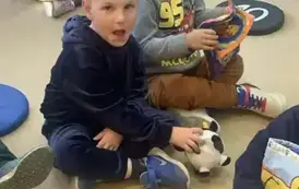 Dw&oacute;ch chłopc&oacute;w na przedzie przegląda pluszowe książeczki, jeden z chłopc&oacute;w ma lekko otwartą buzię. w tle widać inne dzieci.