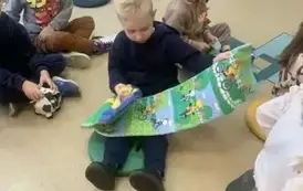 Dzieci siedzą na siedziskach i bawią się pluszowymi książeczkami. Jeden z chłopc&oacute;w rozłożył pluszową książeczkę przeglądając ją.