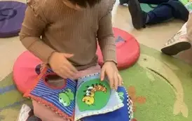 Dziewczynka w ciemnych włosach i beżowej bluzce bawi się pluszową książeczką.