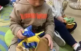 Chłopiec w blond włosach i beżowej bluzie siedzi na dywanie i bawi się pluszową misiową książeczką . Za nim widać inne dzieci bawiące się książeczkami.