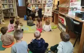 Dzieci siedzą na dywanie i siedziskach i patrzą jak ich kolega stoi przy bibliotekarce i bawi się robiąć zakupy do siateczki korzystając z pluszowej książki przedstawiającej sklep.