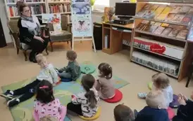 Dzieci siedzą na dywanie i siedziskach patrząc na bibliotekarkę, kt&oacute;ra ma obok na fotelu przygotowane bajeczki do czytania.