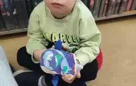 Chłopiec w blond włosach i zielonej bluzie siedzi na siedzisku i bawi się pluszową książeczką.