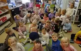 Dzieci siedzą dużą grupą na dywanie, a za nimi na krzesełku siedzi bibliotekarka pokazując okładki dw&oacute;ch książeczek.