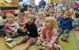 Dzieci siedzą na dywanie i uważnie słuchają bajki, kt&oacute;rą czyta im bibliotekarka.