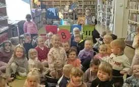 Dzieci siedzą na dywanie i siedziskach, za nimi na krzesełku siedzi bibliotekarka i pokazuje okładki dw&oacute;ch książeczek.