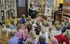 Dzieci dużą grupą siedzą na dywanie i siedziskach, z przodu siedzi bibliotekarka, kt&oacute;ra czyta im bajkę pokazując wnętrze książki. 