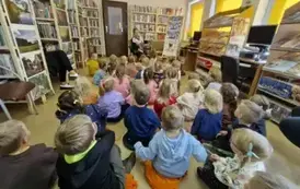 Dzieci dużą grupą siedzą na dywanie i siedziskach, a z przodu bibliotekarka czyta im bajkę pokazując wnętrze książki.