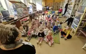 Dzieci dużą grupą siedzą na dywanie i siedziskach, z przodu siedzi bibliotekarka, a z tyłu za dziećmi siedzą ich Panie.