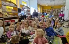 Dzieci dużą grupą siedzą na dywanie i siedziskach słuchając bajki, kt&oacute;rą czyta im bibliotekarka. Za nimi po bokach siedzą ich Panie z Przedszkola.
