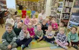 Dzieci siedzą na dywanie, za nimi siedzi ich Pani.