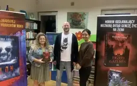 Przy pisarzu stoi kierowniczka biblioteki z nową książką autora, a z prawej stron stoi bibliotekarka. Po bokach stoją rolapy promujące książki pisarza.