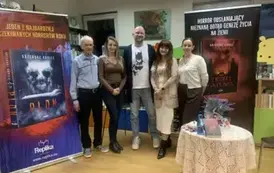 Grzegorz Kopiec stoi z żoną, po bokach stoją jeszcze dwie kobiety i jeden Pan w siwych włosach. Przed nimi stolik, a po bokach rolapy promujące książki pisarza. 