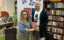 Kierowniczka biblioteki stoi razem z pisarzem trzymając razem najnowszą Jego książkę " Trzeci gatunek". Za nimi stoi rolap z logiem biblioteki.