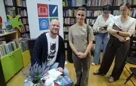 Przy stoliku Grzegorza Kopca stoją czytelnicy z nowymi książkami prosząc o podpis. Pani z przodu i pisarz uśmiechają się.